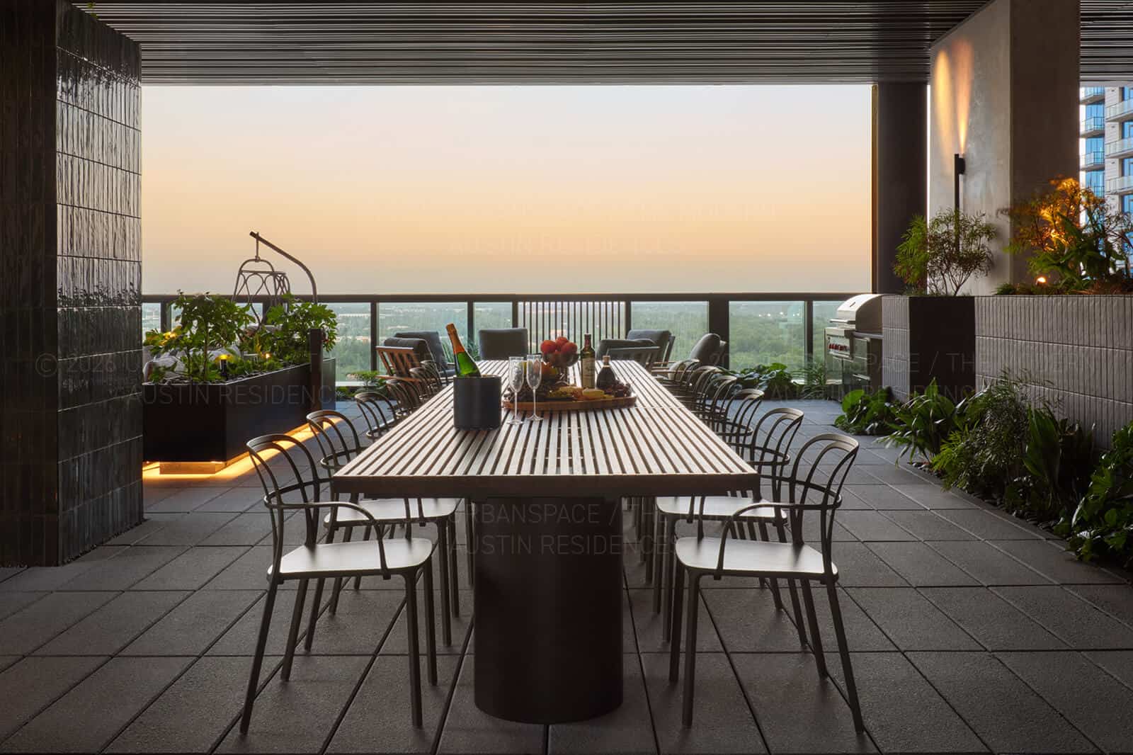 Long dining table on a terrace at sunset, planted troughs and grill beyond